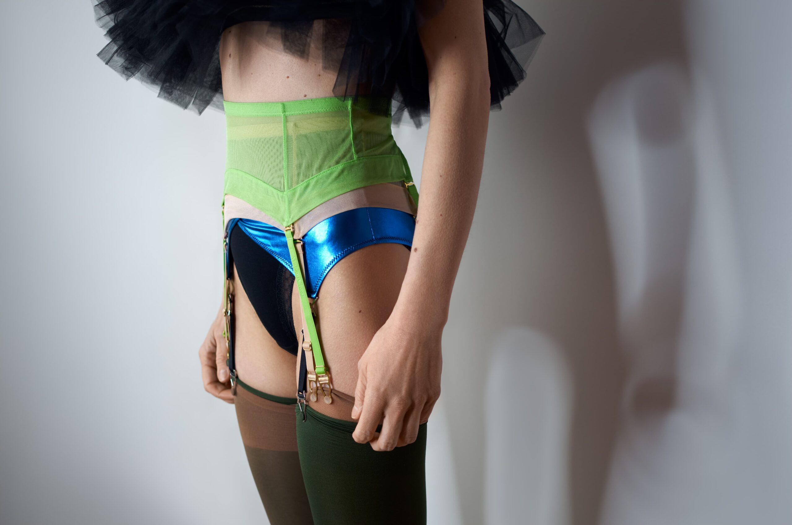Autoportrait partiel d'une personne debout, vêtue de lingerie structurée aux couleurs vives (vert, bleu, noir), montrant le bas du torse et les jambes. Le visage est hors champ. L’éclairage crée une ombre projetée sur un mur blanc.